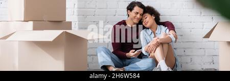 happy multiethnic couple hugging while sitting near carton boxes, banner Stock Photo