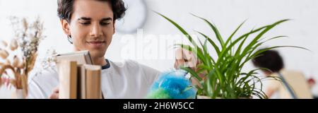 young man doing house cleaning with dust brush near plants and books, banner Stock Photo