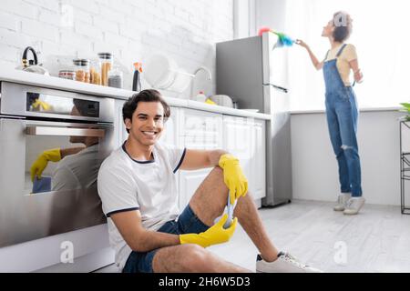 happy young man sitting near kitchen cabinet near curly african american woman cleaning fridge with dust brush in kitchen Stock Photo