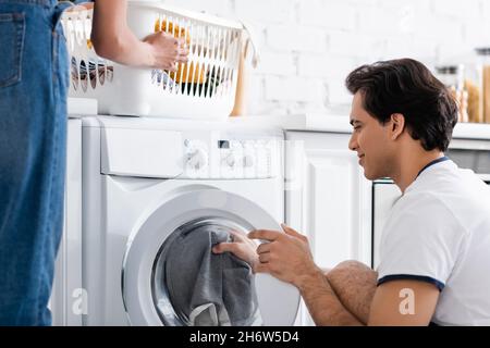 smiling man loading washing machine near african american girlfriend with basket of dirty laundry Stock Photo