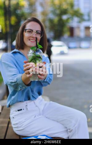 portrait of young elegant business woman in eyeglasses and casual clothes. Woman walking in the city street. Drinking healthy smoothie, reading a book Stock Photo