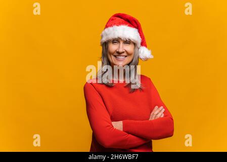 Happy confident senior grey-haired woman wearing red santa hat stands with arms crossed and looking at the camera isolated on yellow. Mature lady promoting new year deal, holiday sale Stock Photo