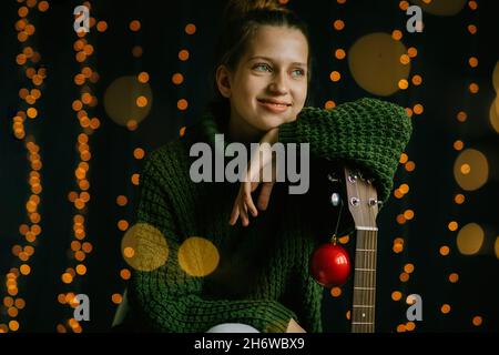 A beautiful teenage girl dreamily looks to the side, smiles broadly, leaning on the neck of a guitar. Christmas decor and home light. Lifestyle, horiz Stock Photo