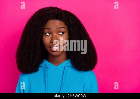 Photo of funny doubtful dark skin woman dressed blue sweatshirt biting lip looking empty space isolated pink color background Stock Photo