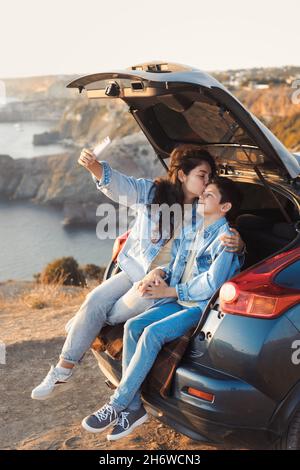 Young mother and her teenage boy in denim fashion clothes sitting in open car trunk, hugging and make selfie Stock Photo