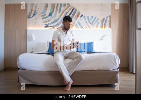 Young attractive businessman sitting on hotel room bed looking at mobile phone. Stock Photo