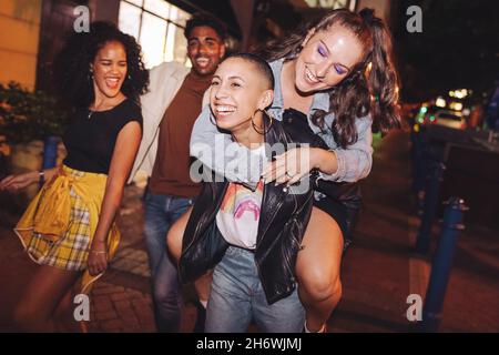 Carefree young woman piggybacking her friend in the city. Girlfriends having a good time while going out with their friends at night. Group of vibrant Stock Photo