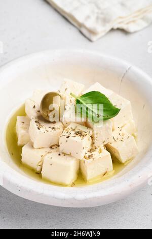 Feta cheese cubes with rosemary, olives and olive oil sauce in white bowl on light gray background. Traditional Greek homemade cheese. Selective focus