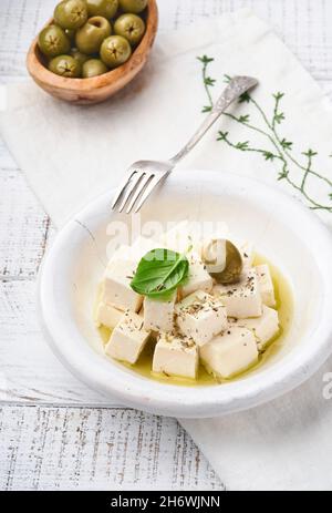 Feta cheese cubes with rosemary, olives and olive oil sauce in white bowl on light gray background. Traditional Greek homemade cheese. Selective focus