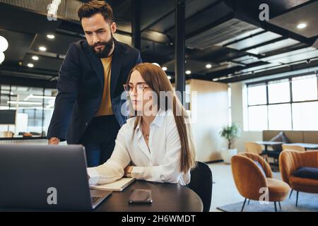 Two businesspeople having a discussion while working with a laptop in an office. Focused entrepreneurs collaborating in a modern workspace. Two young Stock Photo