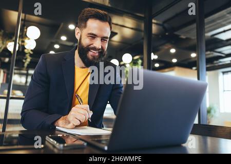 Entrepreneur writing notes during a virtual meeting in an office. Happy young businessman smiling while taking a video call on his laptop. Cheerful bu Stock Photo
