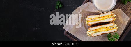 Roasted sandwich cheese and scrambled eggs on paper with coffee glass cup on wooden board. Dark background. Top view, above Stock Photo