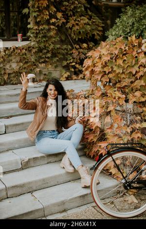 Pretty young woman with mobile phone drink coffee to go at the stairs by the bicycle on autumn day Stock Photo