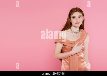 Red haired woman in peach silk dress and sweater looking at camera isolated on pink Stock Photo