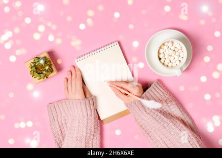 New year, Christmas or holiday wish list concept. Notepad, golden tinsel, and a cup of hot chocolate with marshmallows and female's hands writing Stock Photo