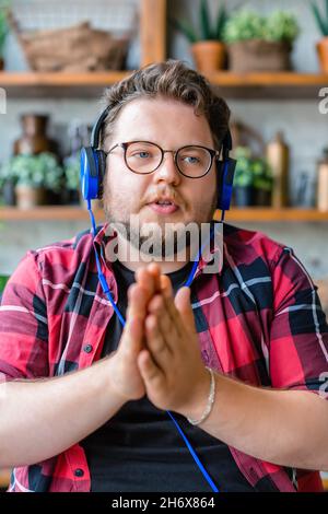 A young bearded man in blue headphones during an online call or online streaming. He looks at the camera and actively gestures during a conversation. Stock Photo