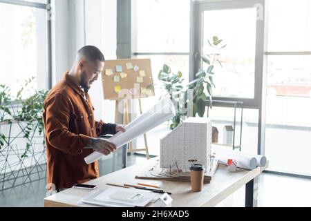 asian architect looking at blueprint while standing near house model and smartphone on desk Stock Photo
