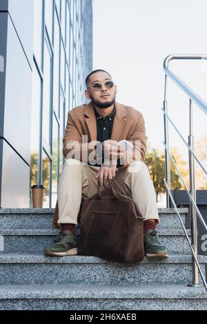 low angle view of asian man in coat and sunglasses sitting on stairs with smartphone and briefcase Stock Photo