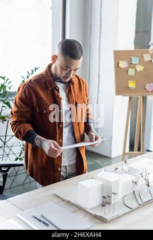 asian architect looking at document while standing near house models on work desk Stock Photo