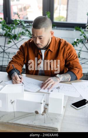 asian architect drawing blueprint while looking at blurred house models near smartphone with blank screen Stock Photo