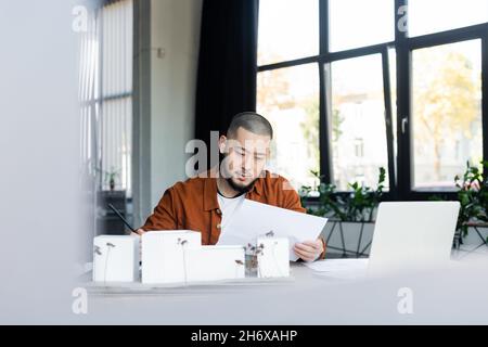 asian architect looking at document while working near blurred laptop and house models Stock Photo