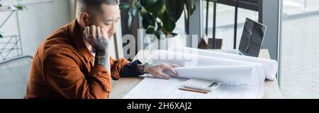 serious asian architect thinking near blueprint while working on project in office, banner Stock Photo