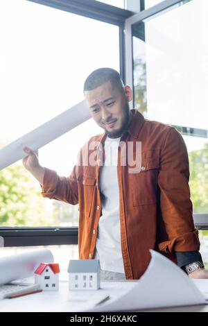 displeased asian architect holding blueprint while standing near house models and papers on work desk Stock Photo