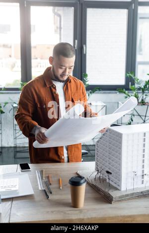 asian engineer looking at blueprint near house model, paper cup and mobile phone on desk Stock Photo