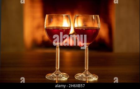 Two red wine glasses on a wooden table, burning fireplace background. Couple drinking, Valentines day celebration dinner or romantic date concept Stock Photo