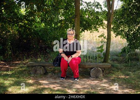 full length view of a mature woman sitting on a wooden park bench on a sunny day in Poland Stock Photo