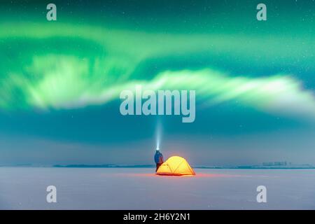 Tourist with flashlight near yellow tent lighted from the inside against the backdrop of incredible starry sky with Aurora borealis. Amazing night landscape. Northern lights in winter field Stock Photo