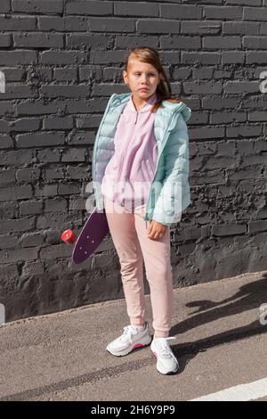 Little girl in colorful clothes stands near gray brick wall with a skateboard in hands Stock Photo