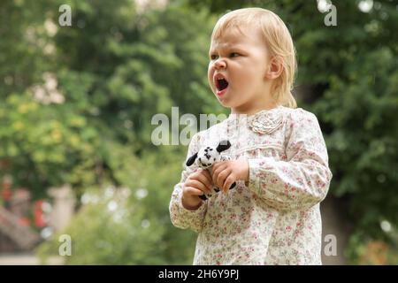 Angry blond baby girl with a toy in hands shouts Stock Photo