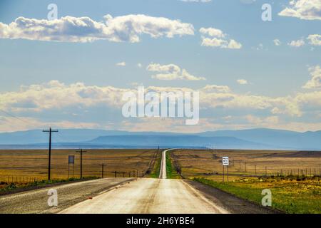 Silver line of two lane highway stretches to horizon with blue layered mountains in the distance and dry fields  and electric lines on each side near Stock Photo