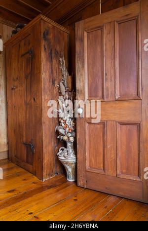 Tall brown stained antique pine wood armoire and ceramic vase with dried plants and flowers next to opened door in principal bedroom inside old home. Stock Photo