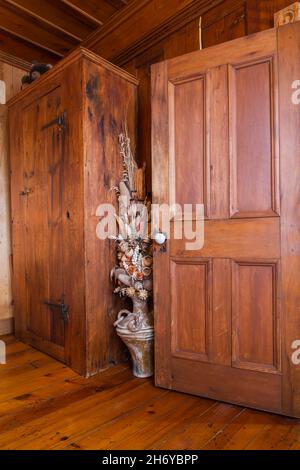 Tall brown stained antique pine wood armoire and ceramic vase with dried plants and flowers next to opened door in principal bedroom inside old home. Stock Photo