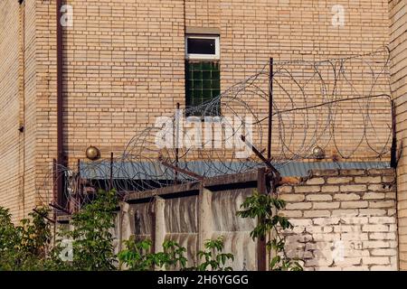 Brick wall of abandoned prison with barred window and fence with rusty barbed wire. Stock Photo