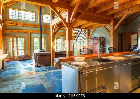 Island with stainless steel countertop and oiled pine wood  bar with black plastic and leather high back chairs in kitchen inside timber frame home Stock Photo