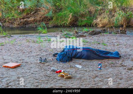 Young homeless person sleeps in sleeping bag under highway overpass on sand bar of East Plum Creek, Castle Rock Colorado USA. Photo taken in October. Stock Photo