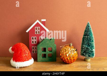 Winter christmas concept. Santa claus hat, house model, bauble, tree on wooden table. red brown background Stock Photo