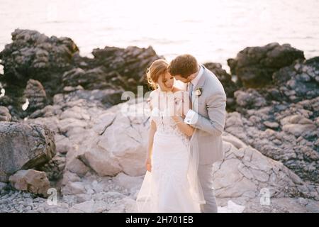 Groom leaned over bride, hugging her shoulders on the rocky seashore Stock Photo