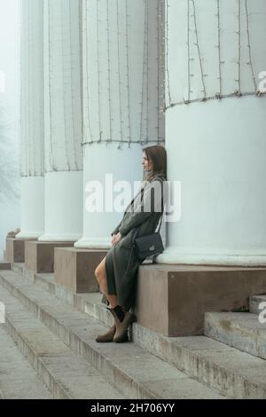 Thoughtful beautiful young woman in green coat standing near white columns. Girl in city Stock Photo