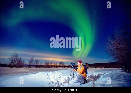 Concept photo tour to arctic travel, photographer man with camera and tripod photographs aurora borealis, northern lights green. Stock Photo