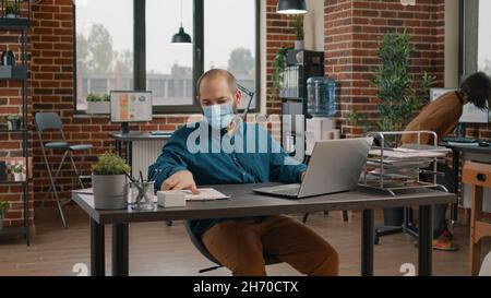Employee checking data rate charts on papers and laptop, looking at display and wearing face mask. Business man using technology to plan project for presentation and startup meeting Stock Photo