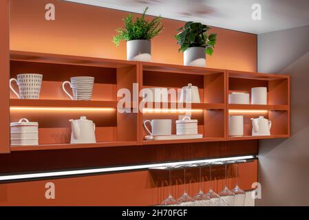 Decorative display of white themed ceramics on a fitted wooden kitchen shelf mounted to a wall with a rack of clean wineglasses in an interior decor a