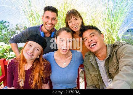 Group of cheerful multiethnic male and female friends in casual clothes smiling and looking at camera while taking selfie in green garden on sunny day Stock Photo