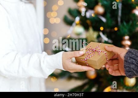 Cropped shot of hands and giftbox, man gives his girlfriend a present tied with white ribbon over blurred christmas tree background.  Stock Photo