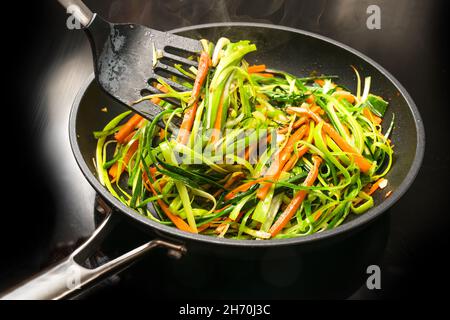 Cooking healthy vegetable noodles from carrot julienne and green leek strips in a black frying pan, for a vegetarian meal, selected focus, narrow dept Stock Photo