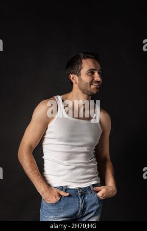 Portrait of young handsome bearded sad man in white undershirt and jeans posing isolated on dark background. Stock Photo