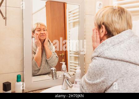 Senior woman in bathrobe touching her soft face skin, looking in mirror at home Stock Photo